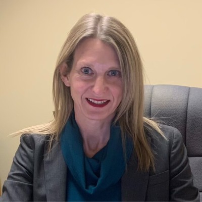 Portrait of a smiling woman with blonde hair, wearing a dark blazer and teal scarf, seated in an office chair.