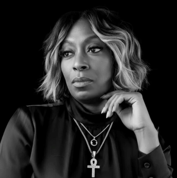 Tiffany Pringle - NAMI Black-and-white portrait of a woman with short wavy hair, wearing a black turtleneck and layered necklaces with an Egyptian ankh pendant, against a black background.