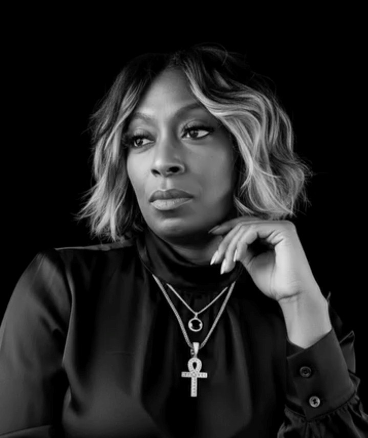 Black-and-white portrait of a woman with short wavy hair, wearing a black turtleneck and layered necklaces with an Egyptian ankh pendant, against a black background.