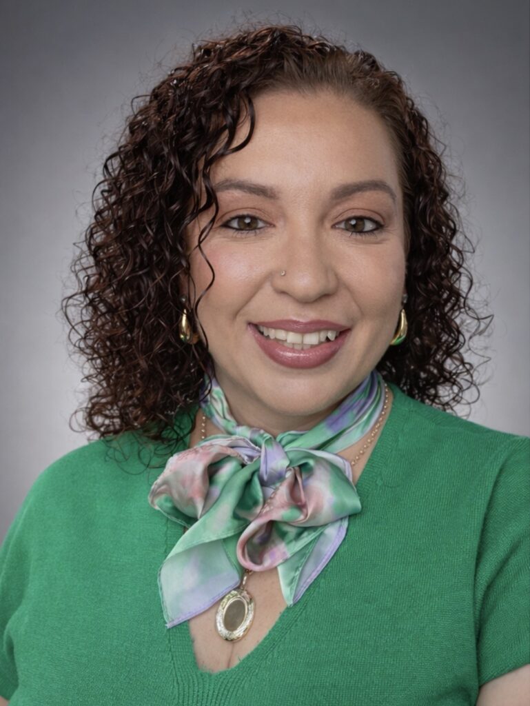 Portrait of a smiling woman with curly brown hair, wearing a green sweater and a pastel scarf with a gold pendant necklace.